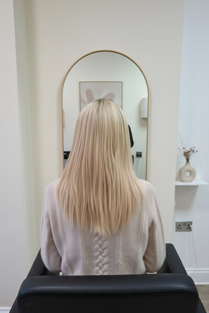 A person with long, straight blonde hair sits in a salon chair, viewed from behind. The person is wearing a cream-colored cable-knit sweater, and a salon mirror and counter are visible in the background.